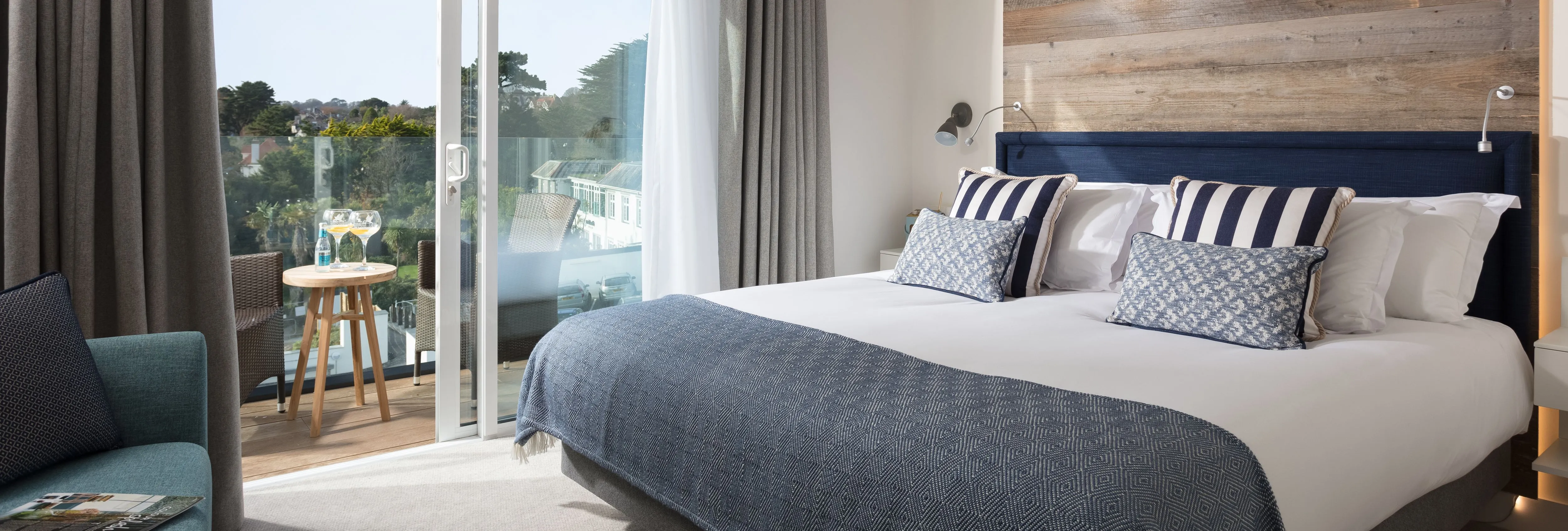 Modern beach house room with large bed, wooden accent wall, and terrace overlooking greenery.
