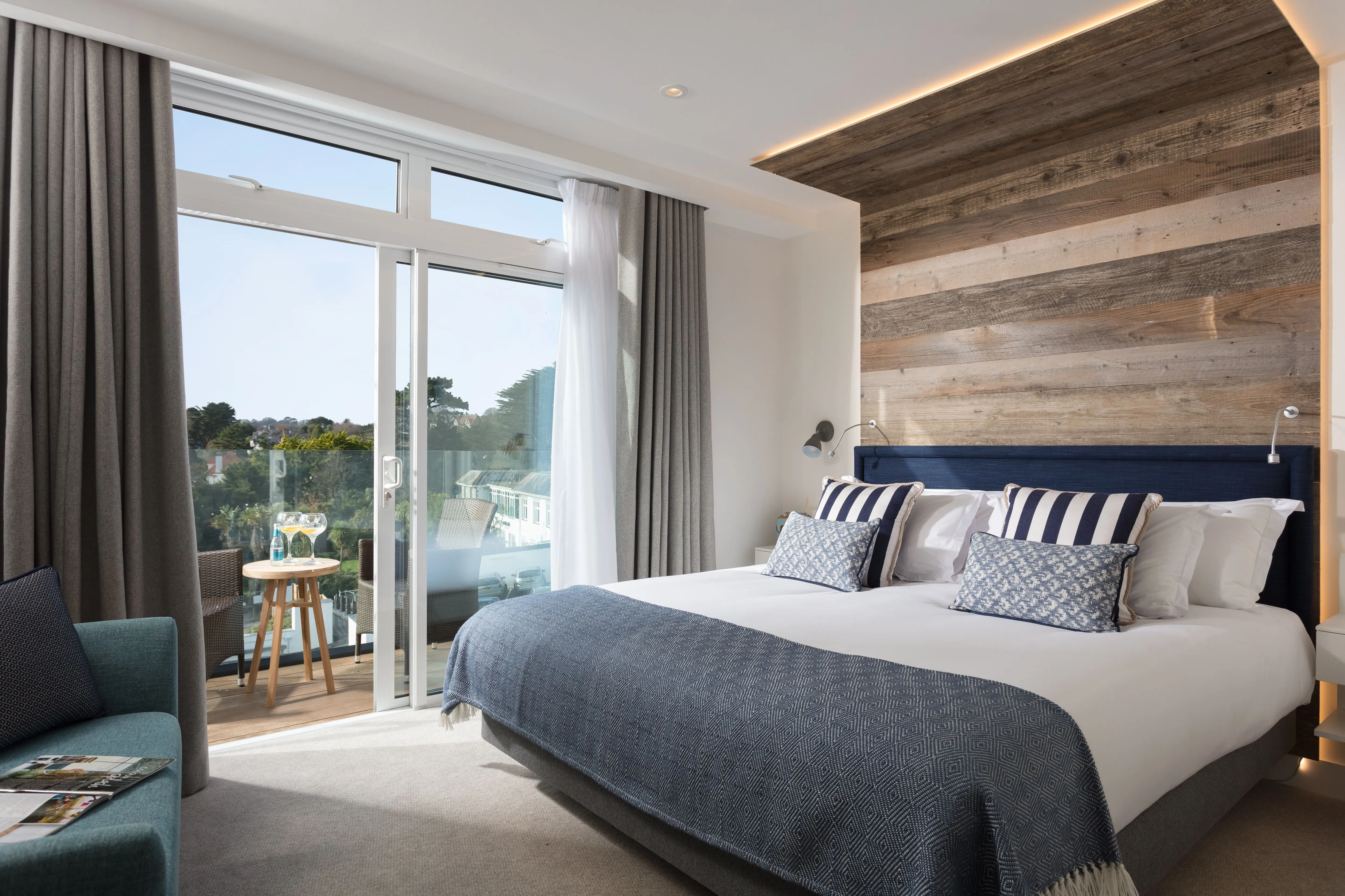 Modern beach house room with large bed, wooden accent wall, and terrace overlooking greenery.