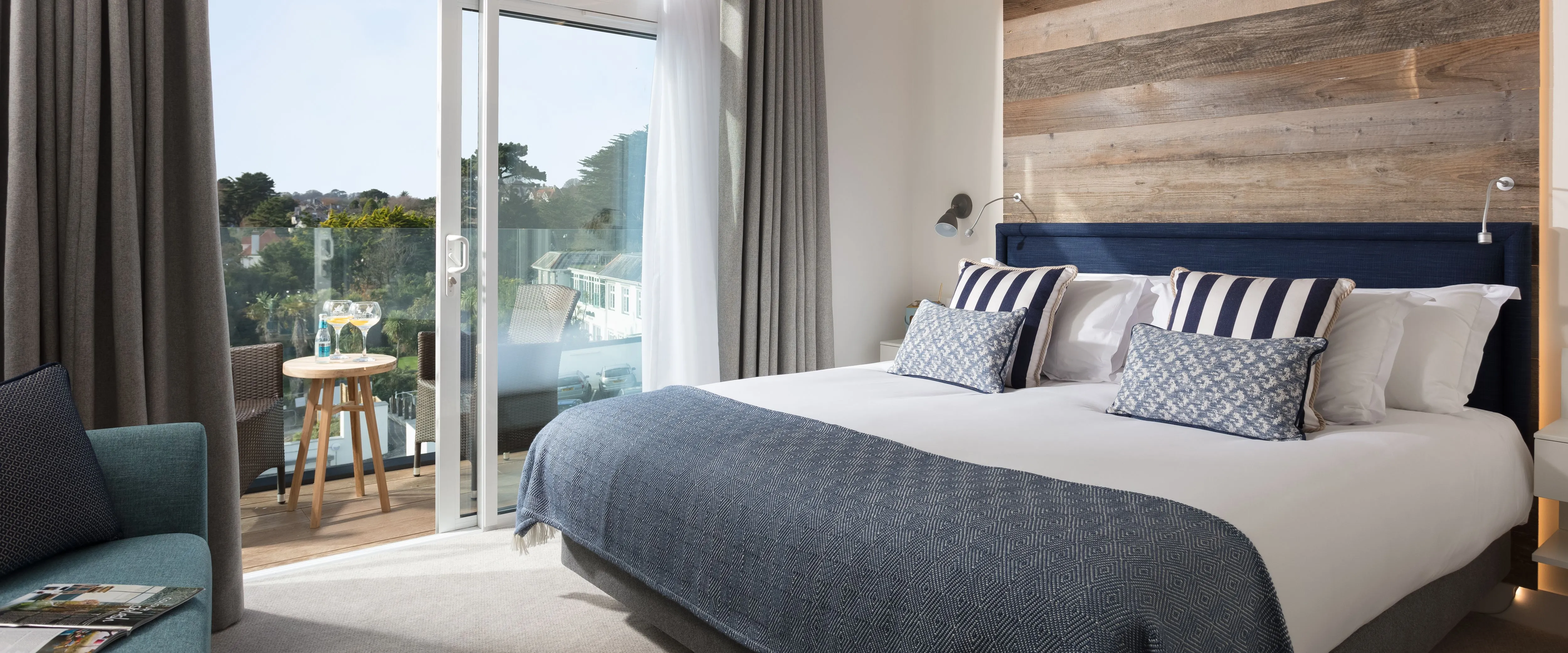 Modern beach house room with large bed, wooden accent wall, and terrace overlooking greenery.