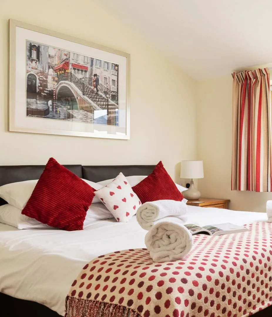 Cosy bedroom with a double bed, red cushions, folded towels and a large window with striped curtains