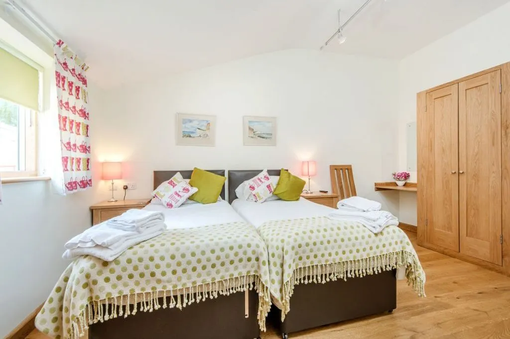 Bright twin bedroom with two single beds, green cushions, wooden furniture and a window with patterned curtains.