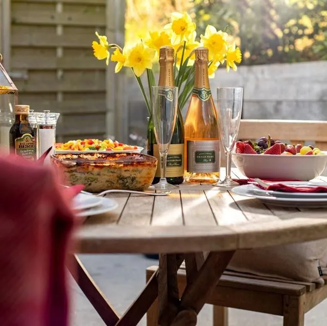 Outdoor garden dining table set with sparkling wine, daffodils, fruit bowl and prepared dishes in warm evening light