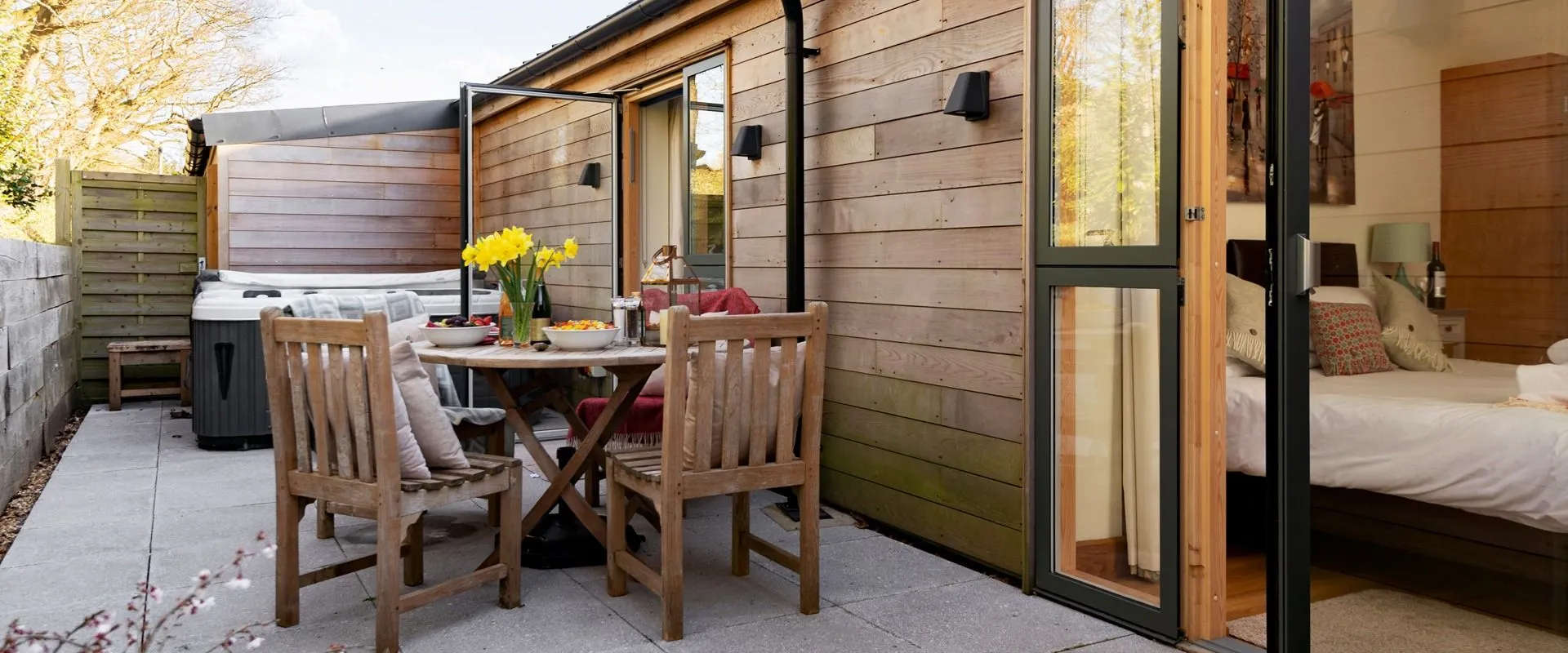 Wood-clad holiday lodge patio with outdoor dining table, chairs and a hot tub beside open doors to a bedroom