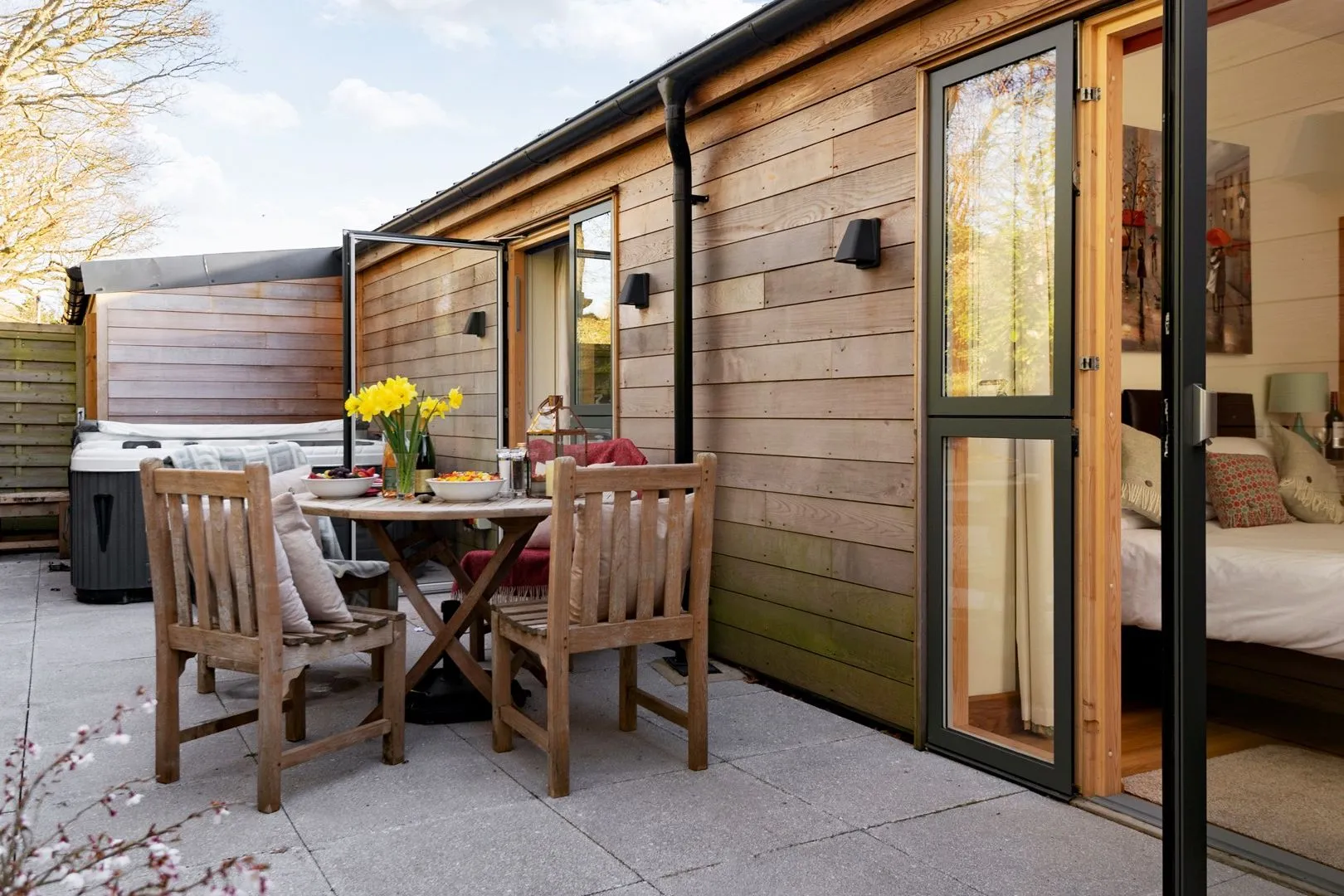 Wood-clad holiday lodge patio with outdoor dining table, chairs and a hot tub beside open doors to a bedroom