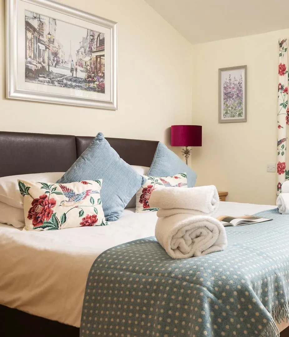 Beautifully decorated bedroom with a double bed, floral cushions and curtains, folded towels, and glass doors opening onto a small patio garden.