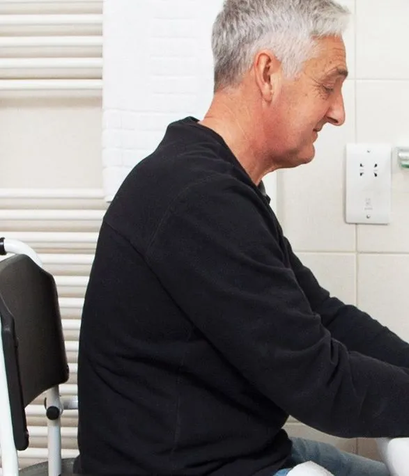 Older man seated at a bathroom sink washing his hands beside a mobility aid chair