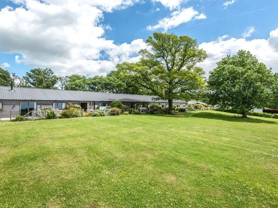 Single-storey countryside property with a large lawn, mature trees and a bright blue sky with scattered clouds.