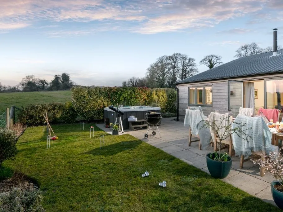 Patio garden beside a modern countryside lodge with outdoor dining, hot tub, barbecue and lawn games at dusk