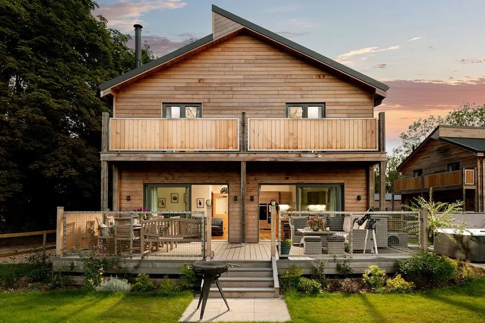 Modern timber-clad house at dusk with two balconies, a decked terrace and warm interior lighting
