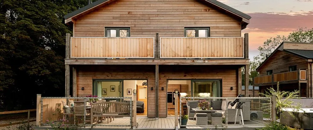 Modern timber-clad house at dusk with two balconies, a decked terrace and warm interior lighting