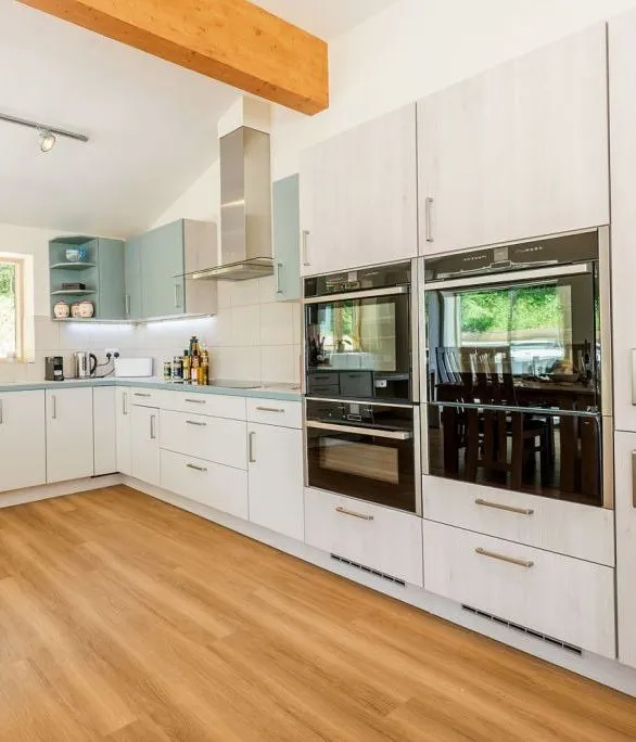 Spacious modern kitchen with white and pale blue cabinets, wooden flooring, integrated double ovens and a large window