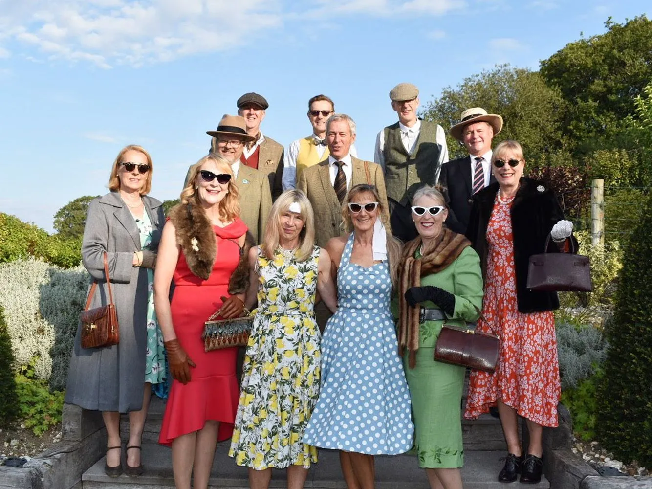 A group of guests dressed in vintage-inspired outfits and hats pose together outdoors in a garden under a blue sky, ready for Goodwood Revival.