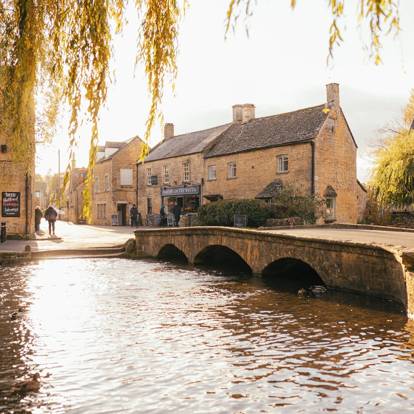 Golden-stone village buildings beside a river with a low stone bridge and overhanging willow branches in warm afternoon light.