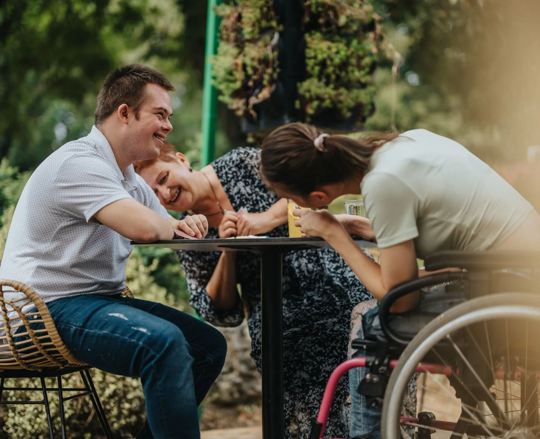 Three people laughing together at an outdoor café table, including a young woman in a wheelchair and a young man with Down syndrome.