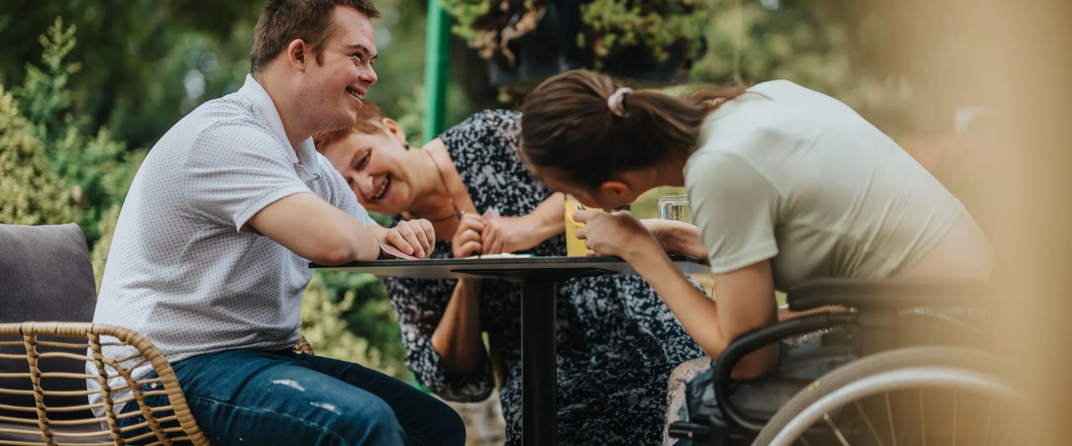 Three people laughing together at an outdoor café table, including a young woman in a wheelchair and a young man with Down syndrome.