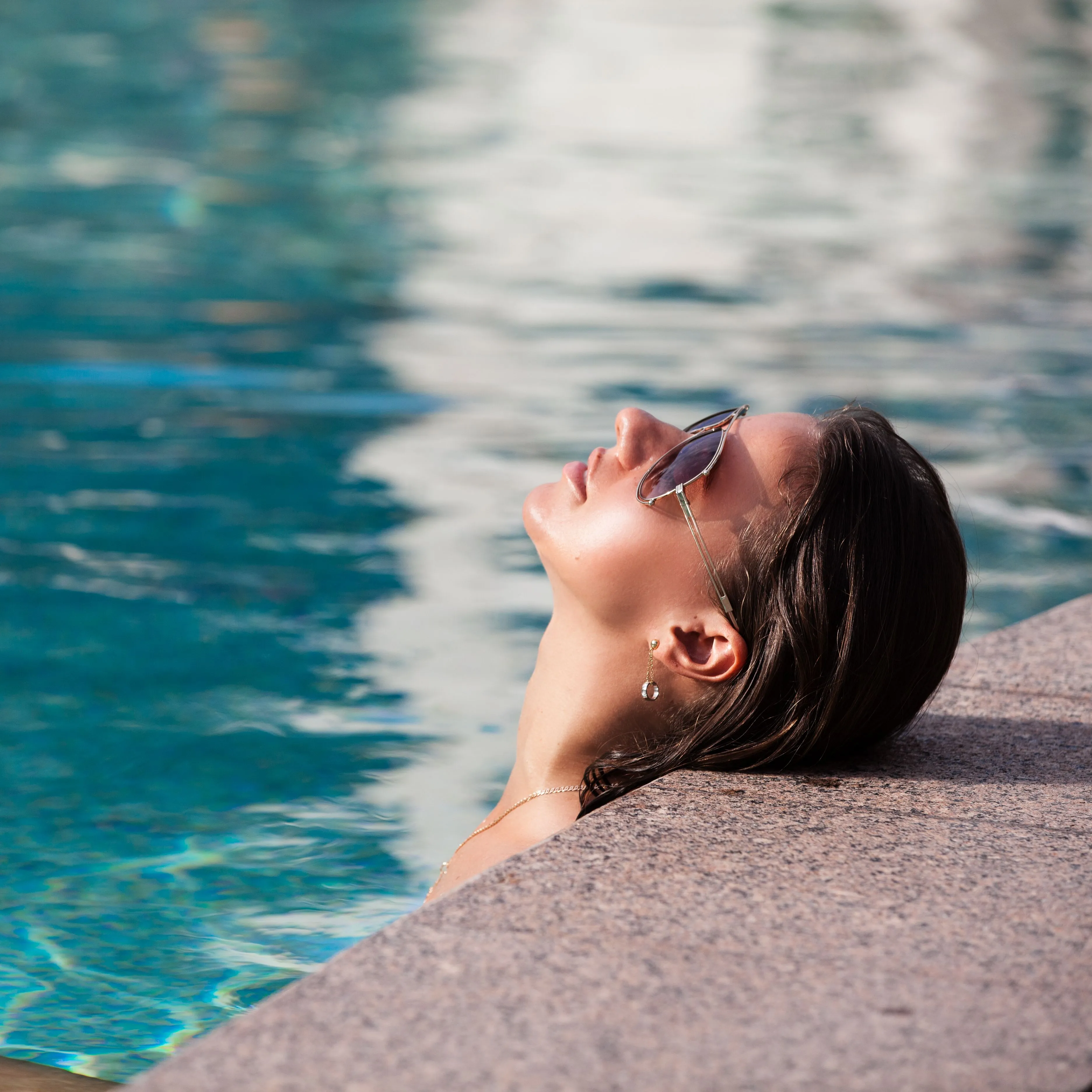 Woman wearing sunglasses relaxing at the edge of a swimming pool in sunlight
