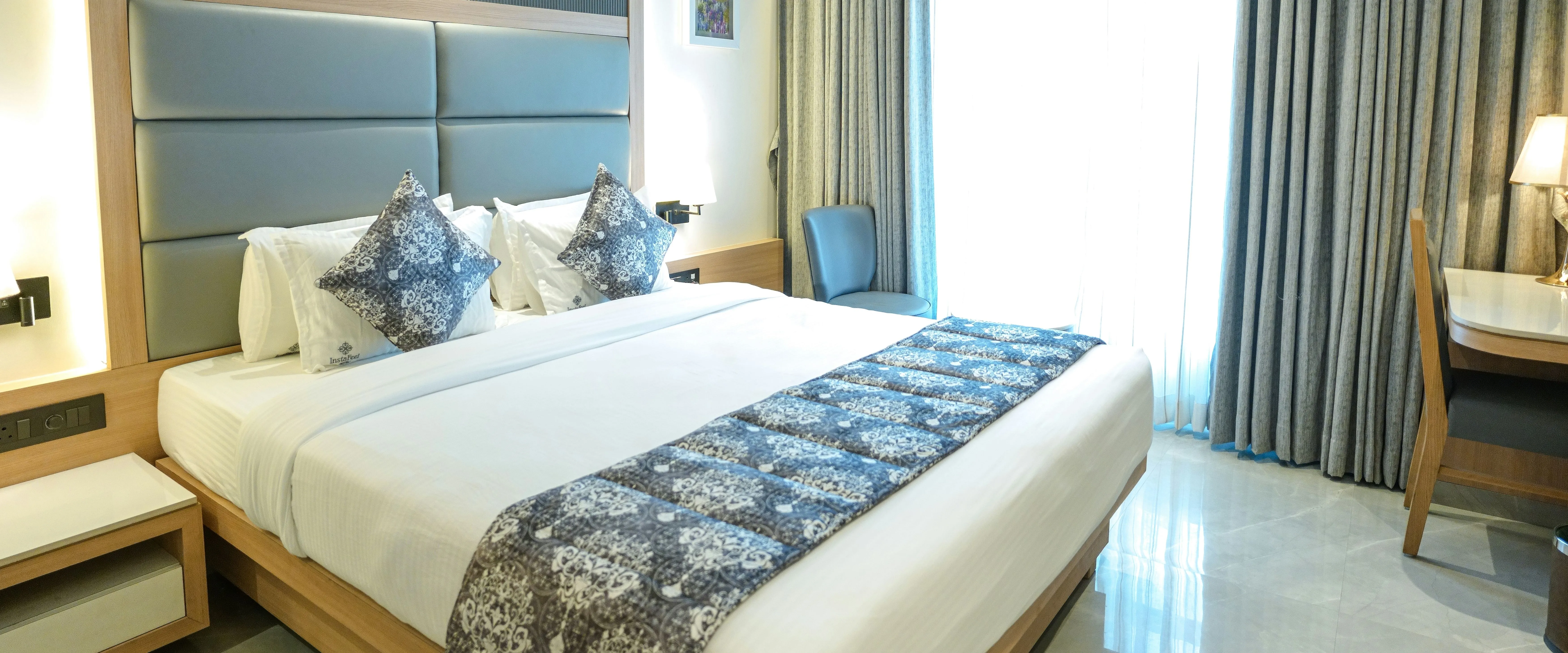 Modern hotel bedroom with a large double bed, grey upholstered headboard and floor-length curtains
