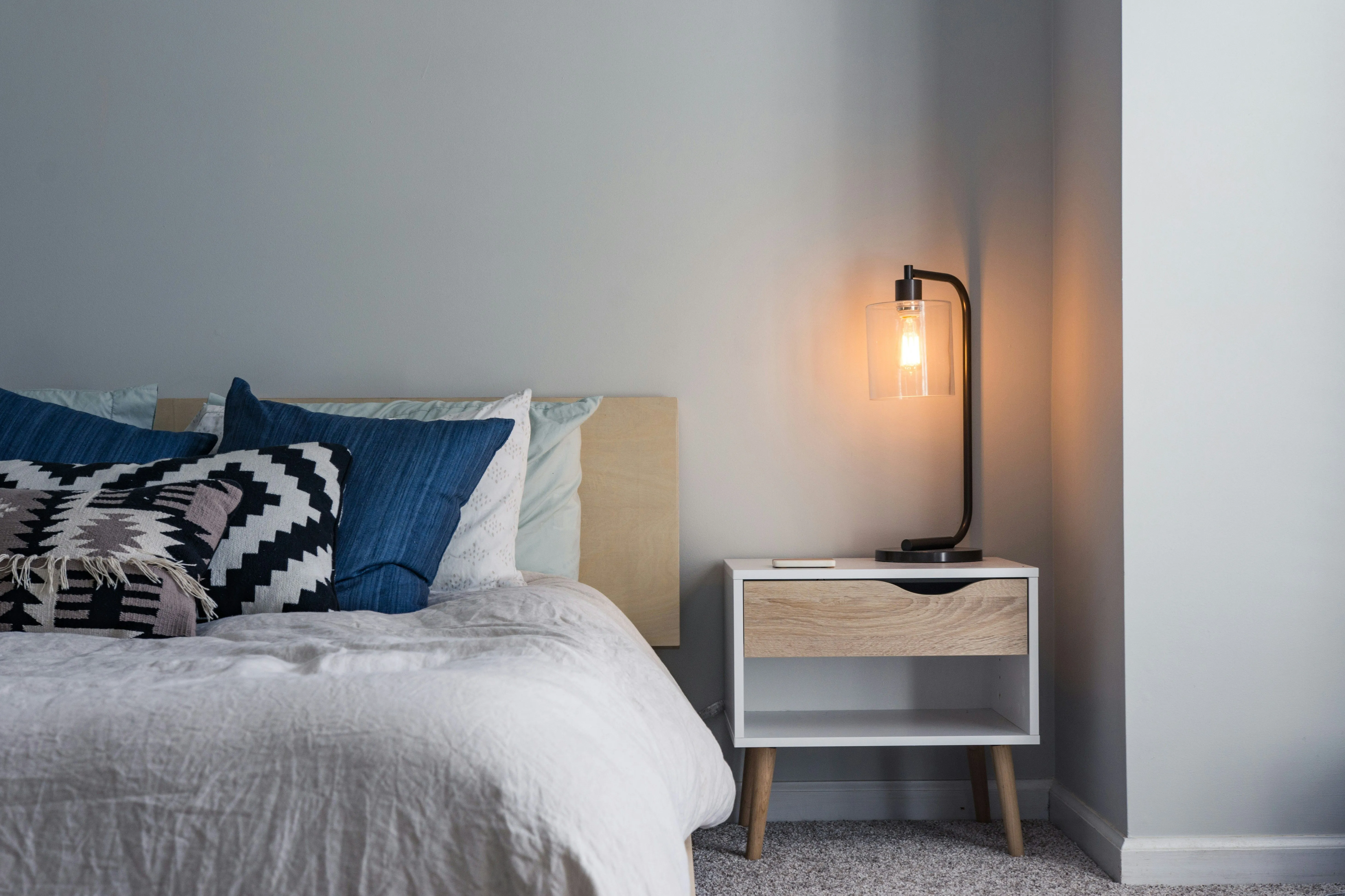 Modern bedroom with white bedding, blue cushions and a lit bedside lamp on a small wooden nightstand
