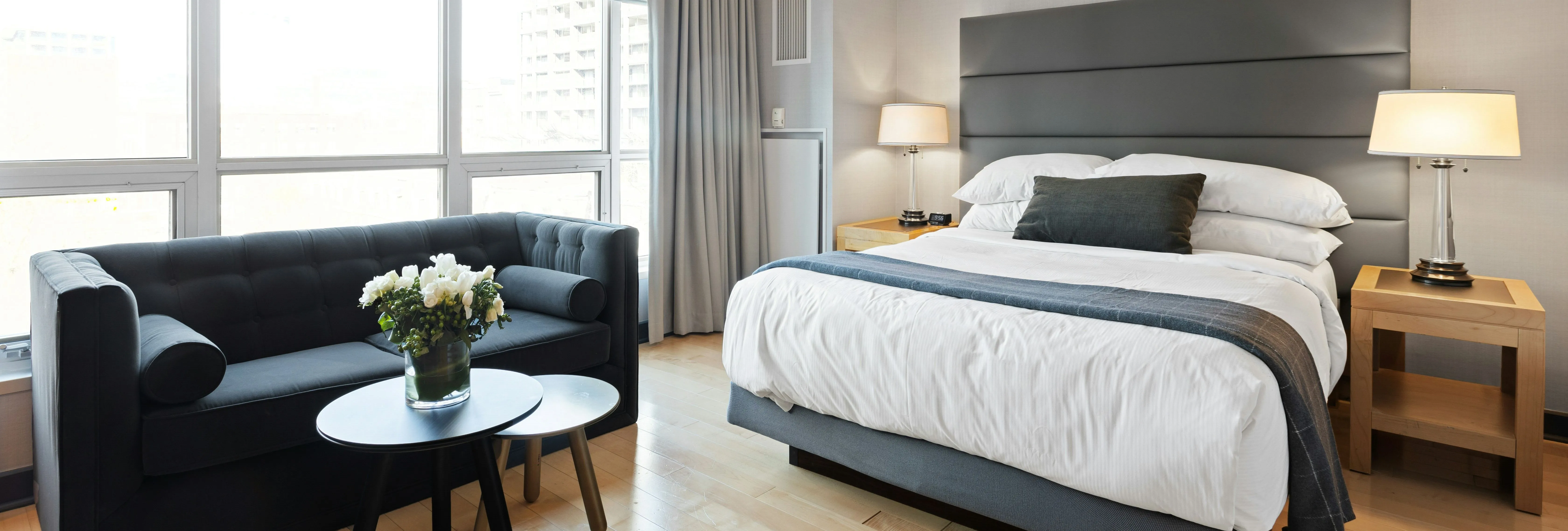 Modern hotel bedroom with a double bed, bedside lamps, large windows and a dark sofa beside a small table with flowers