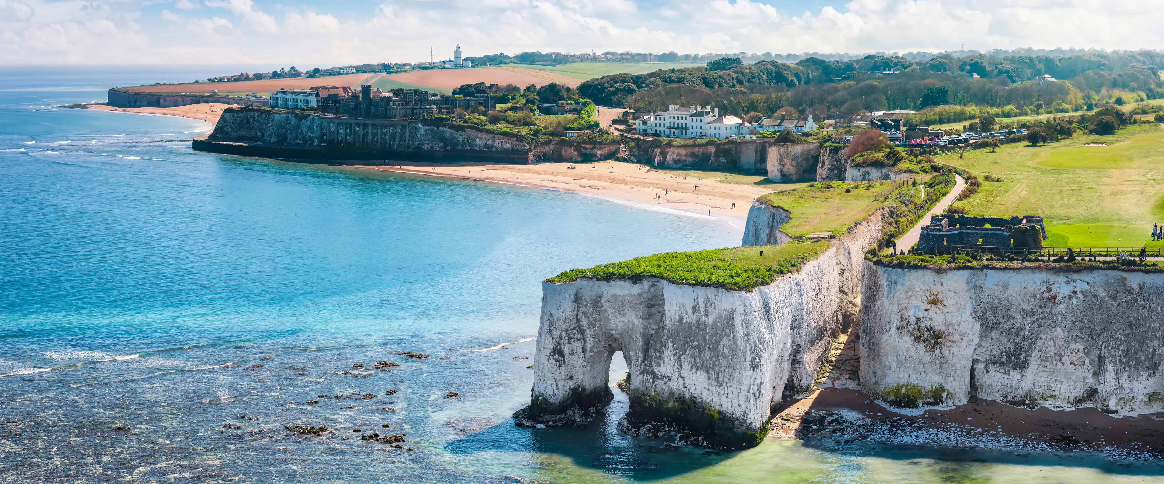 White chalk cliffs and a sea arch overlooking a sandy bay with turquoise water under a bright blue sky.