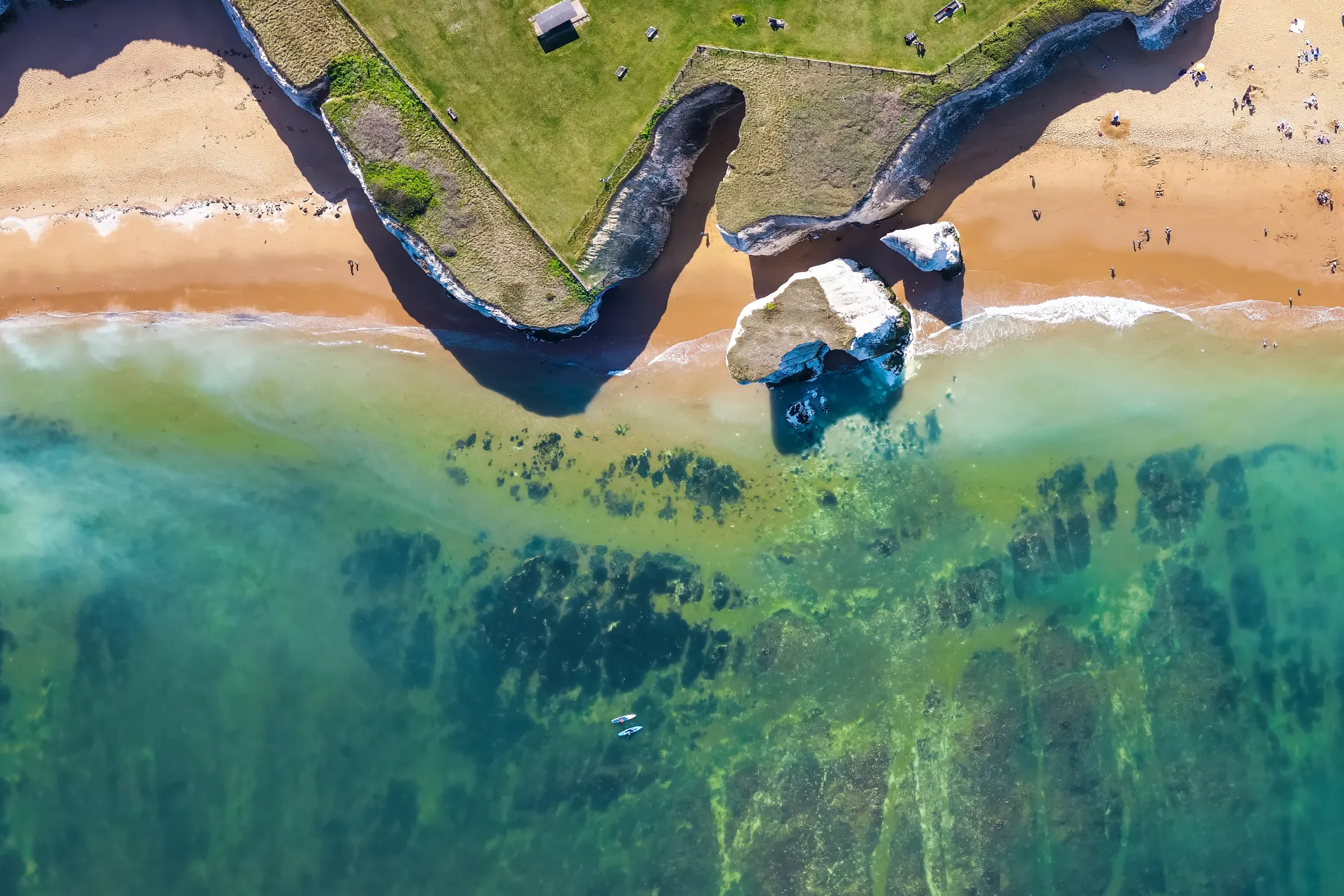 Aerial view of a sandy beach bordered by grassy clifftops, with turquoise sea, rock formations and people scattered along the shore.