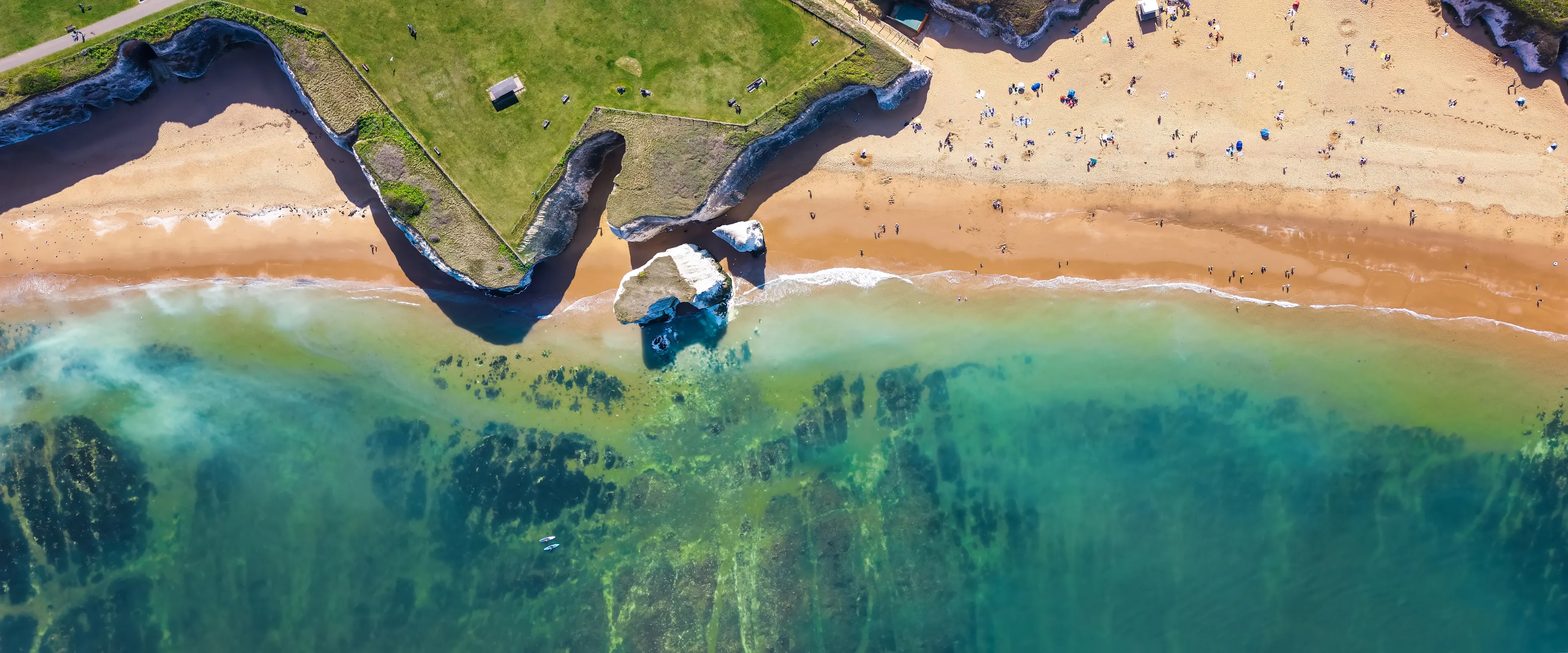 Aerial view of a sandy beach bordered by grassy clifftops, with turquoise sea, rock formations and people scattered along the shore.