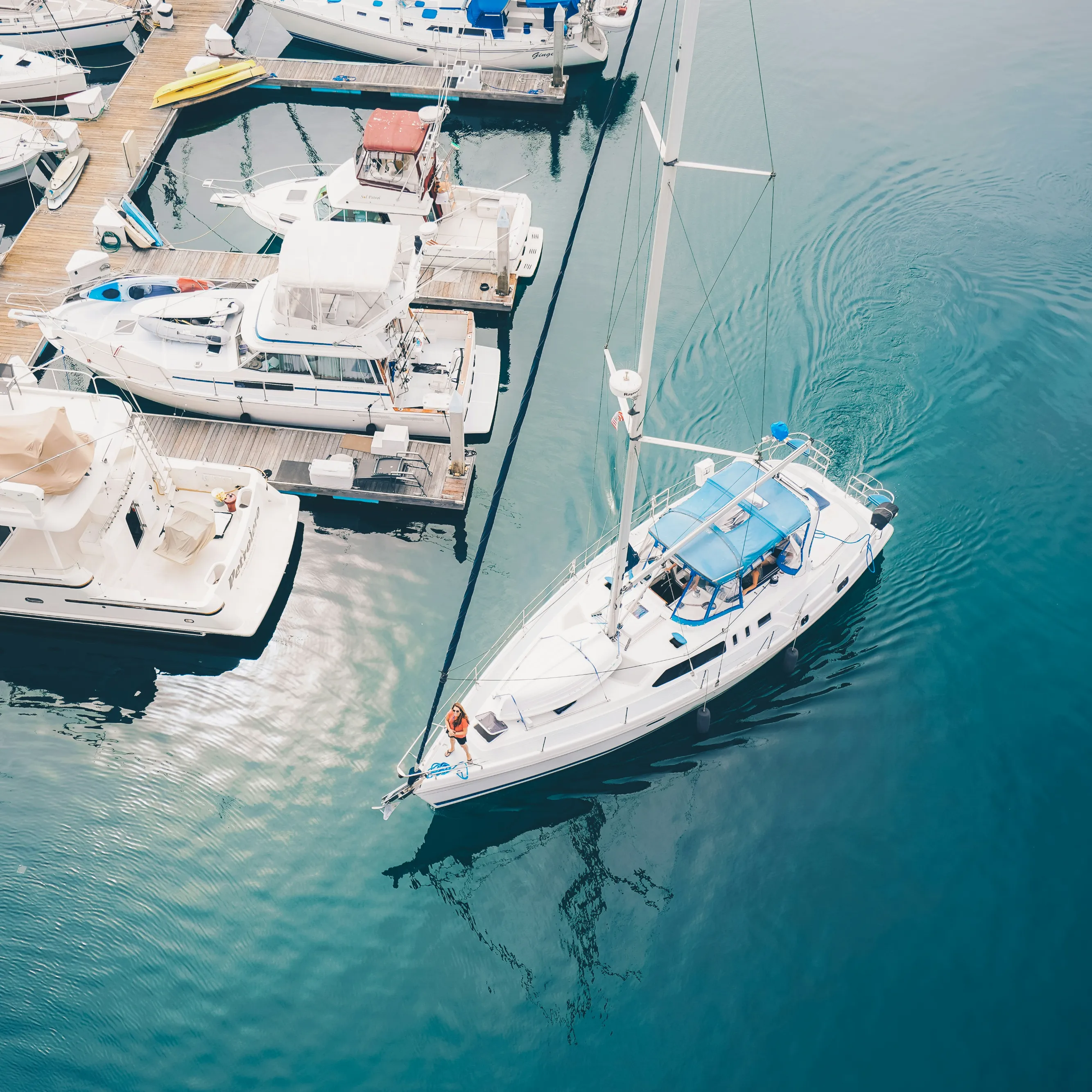 Aerial view of yachts and sailboats moored at a marina, with one white sailing boat moving through calm blue water.