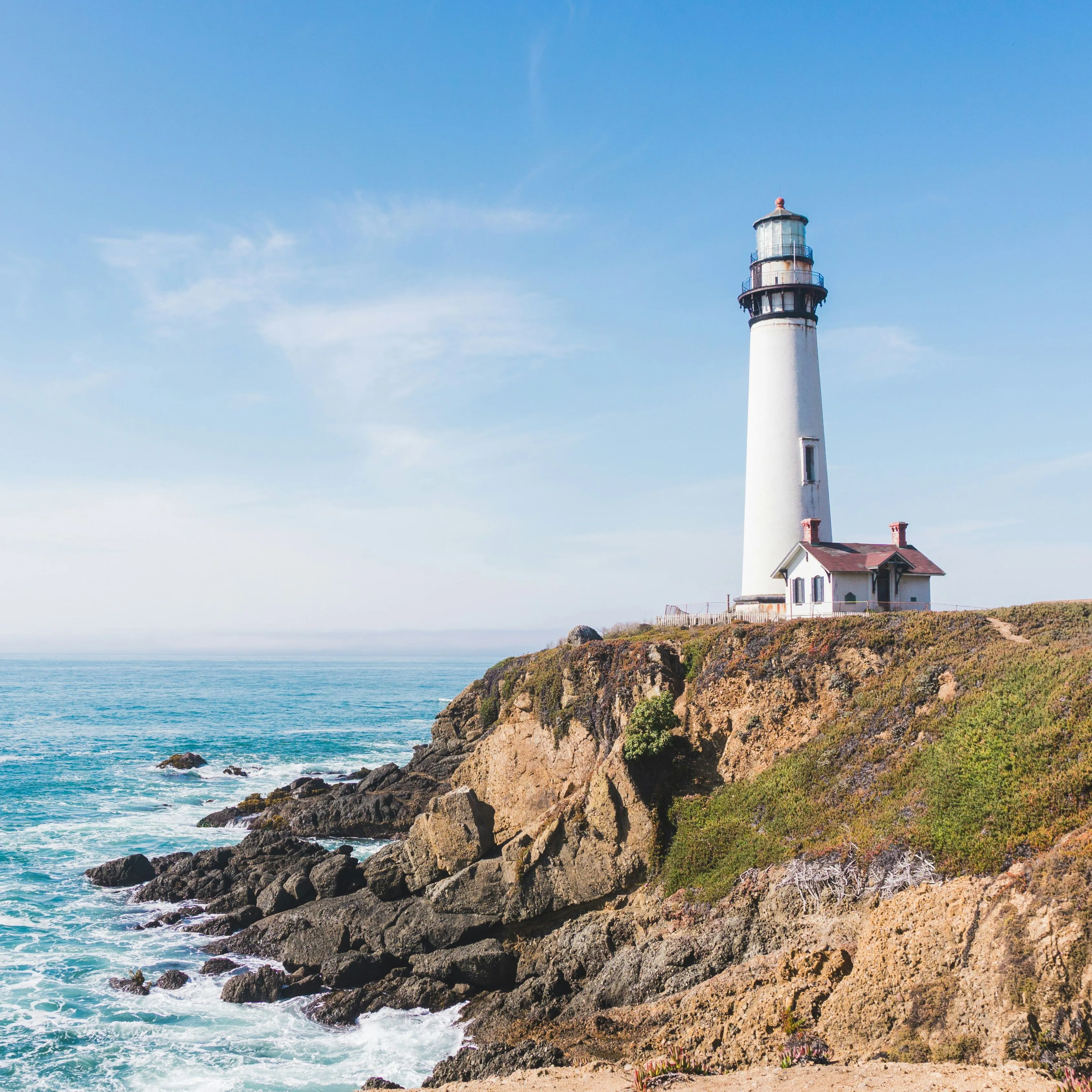 White lighthouse on a rocky cliff overlooking the sea under a clear blue sky