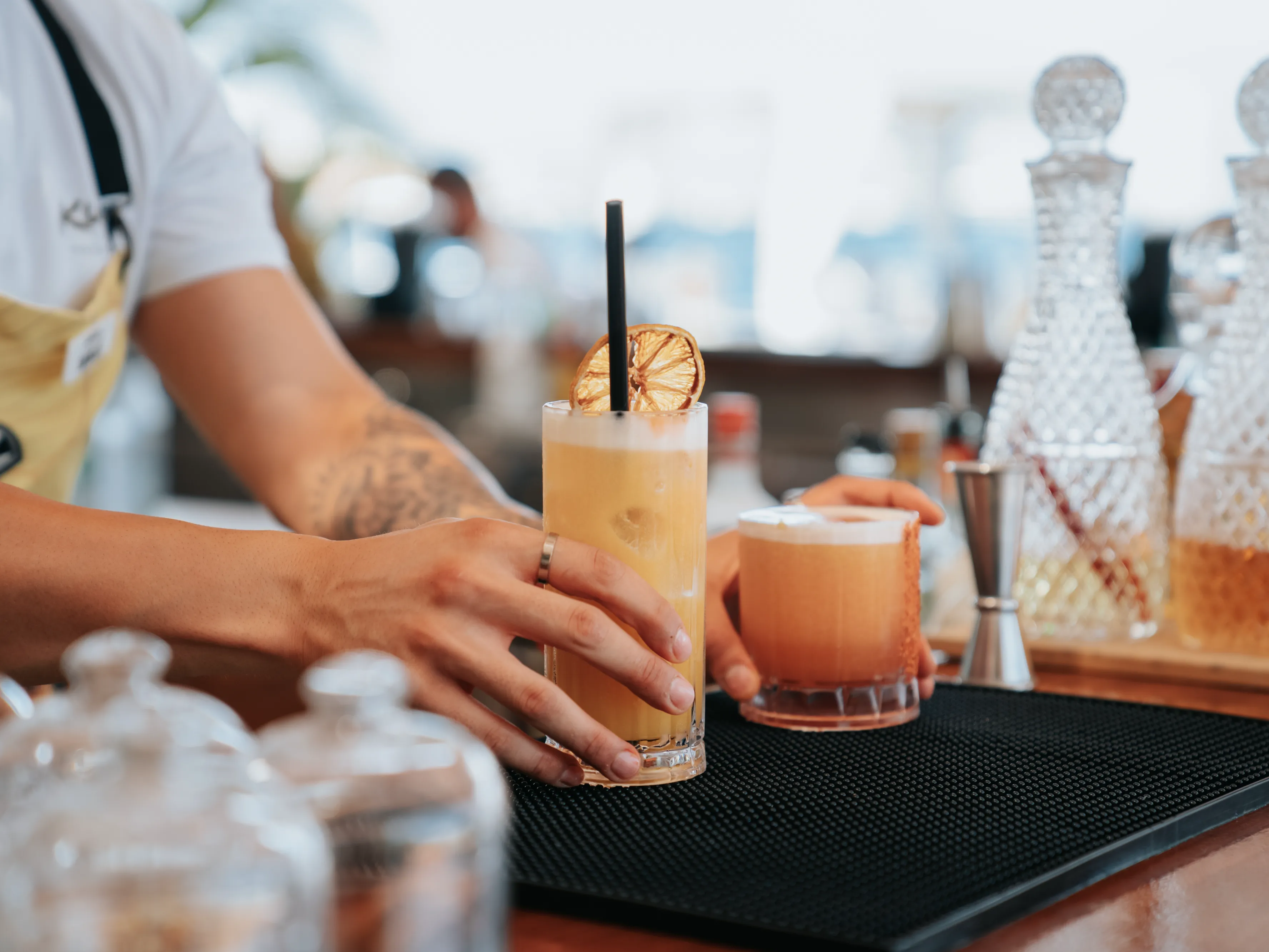 Bartender placing two frothy cocktails on a bar counter, one garnished with a dried citrus slice.