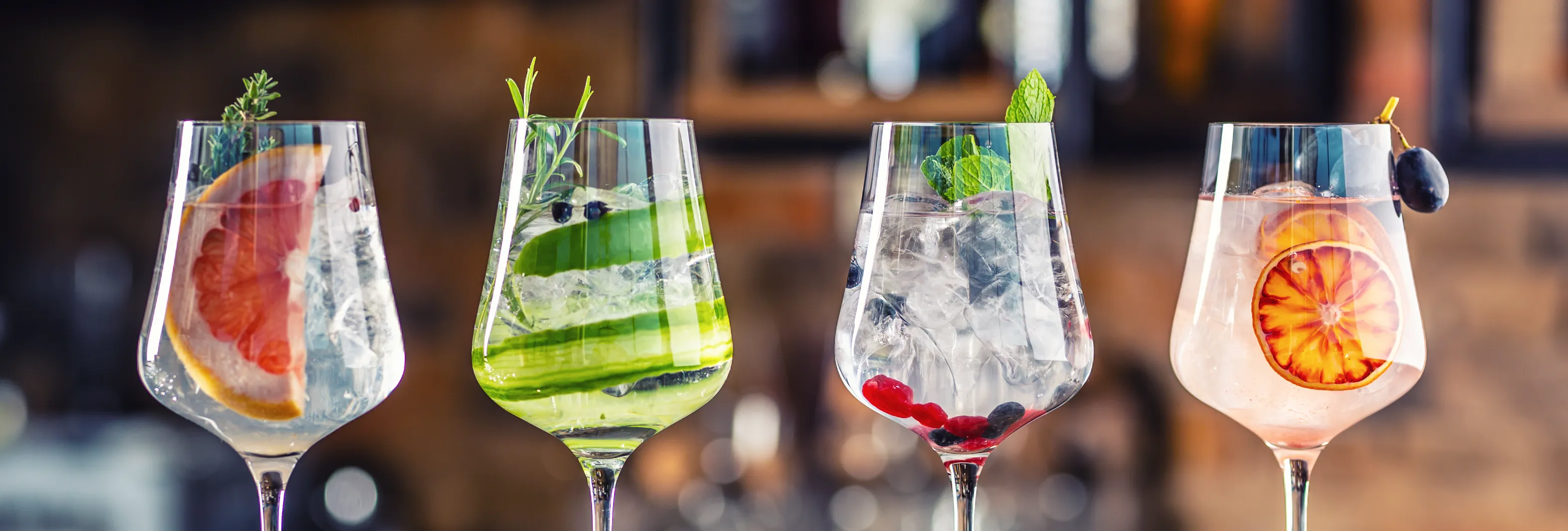 Four elegant cocktails in stemmed glasses on a wooden bar, garnished with grapefruit, cucumber, mint and dried orange.