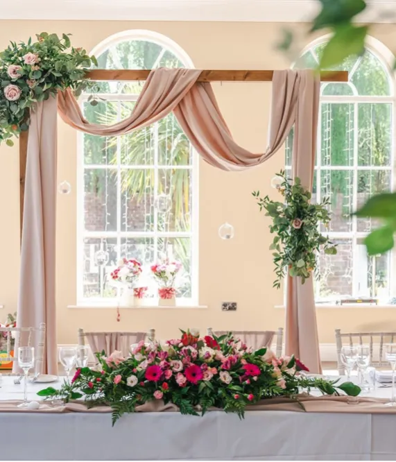 Wedding top table with floral centrepiece and draped ceremony arch in a bright reception room