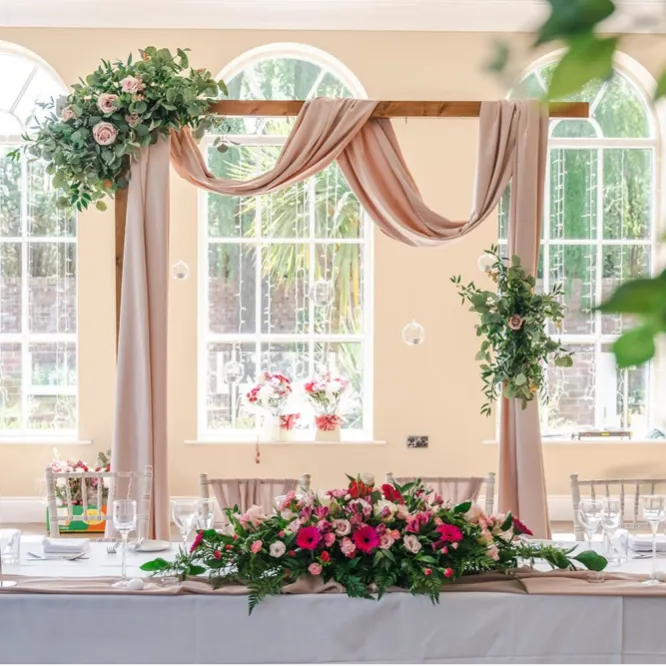 Wedding top table with floral centrepiece and draped ceremony arch in a bright reception room