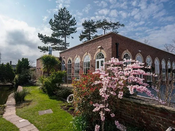 Brick building with arched windows beside a landscaped garden and pond, with pink blossom in the foreground