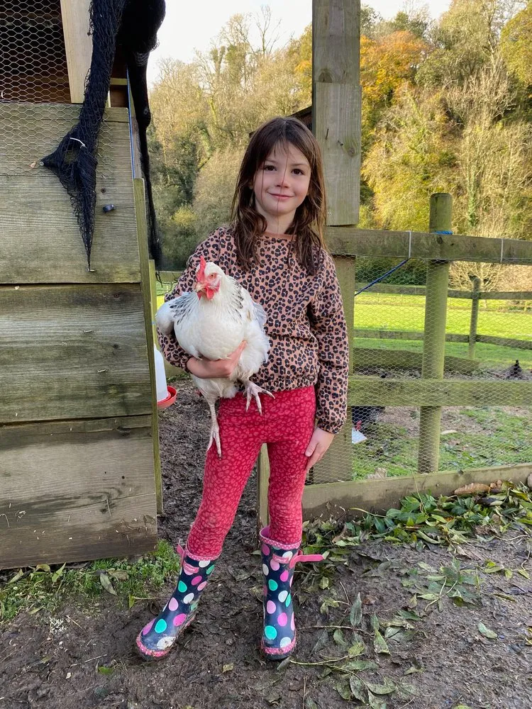A young girl in a leopard-print sweater, red leggings, and colorful polka-dot rain boots stands in a muddy farmyard, smiling while holding a white chicken.