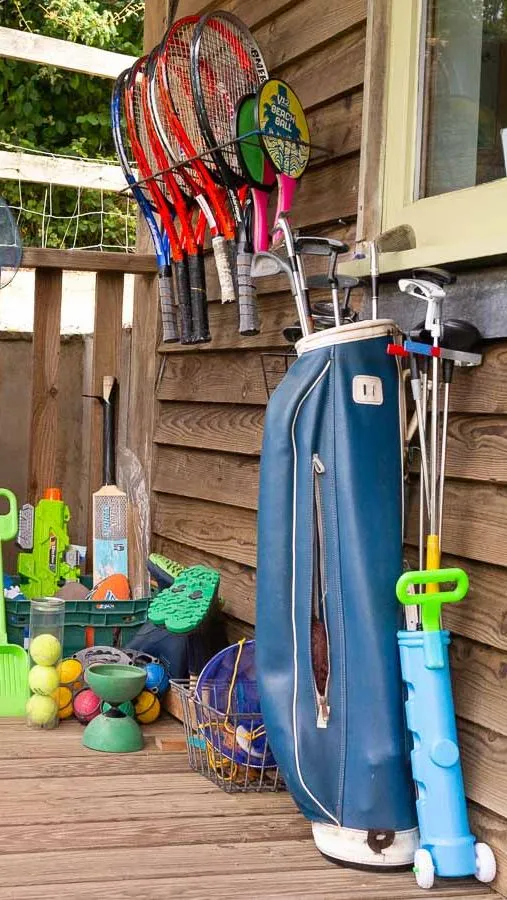 An outdoor storage area with sports and beach equipment, including tennis rackets, golf clubs, buckets, shovels, and inflatable toys, neatly arranged against a wooden wall.