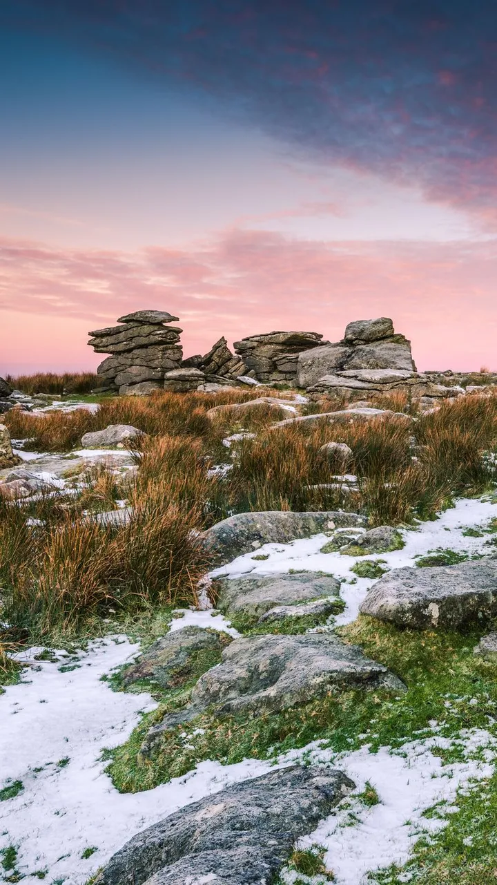 A rugged moorland landscape with weathered rock formations, patches of snow, and tall grasses beneath a vibrant pink and blue sunset sky.