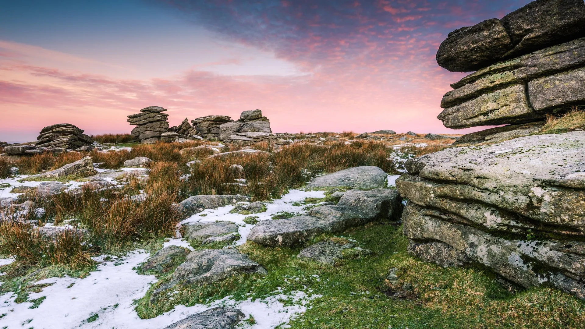 A rugged moorland landscape with weathered rock formations, patches of snow, and tall grasses beneath a vibrant pink and blue sunset sky.