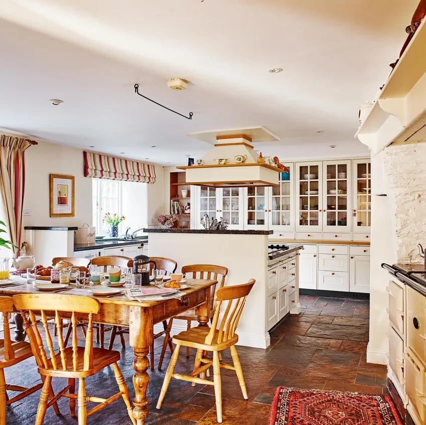 A bright, country-style kitchen with a large wooden dining table set for breakfast, cream cabinetry, an Aga stove, and rustic accents, bathed in natural light.
