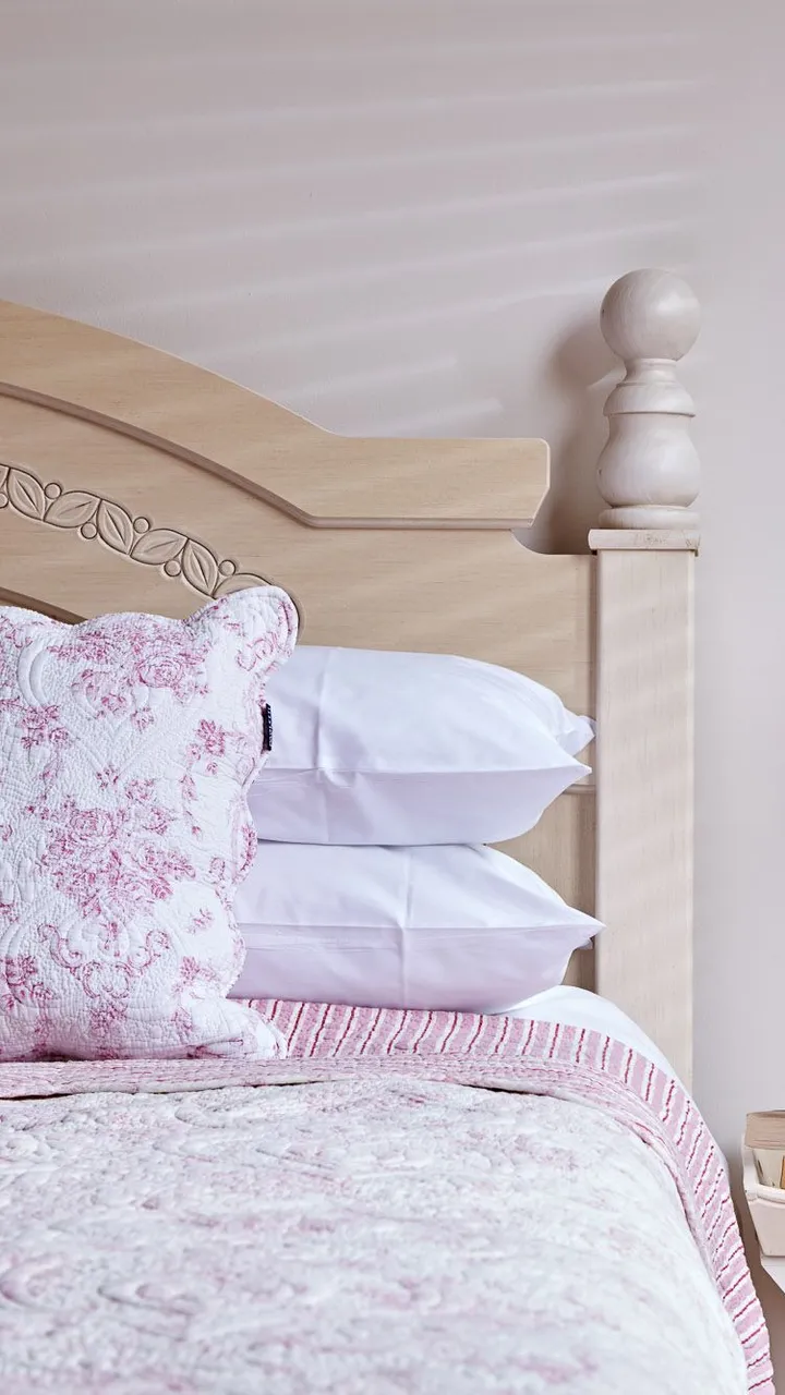 A cozy bedroom with a light wooden bed, pink floral bedding, and a matching cushion. A bedside table holds books, fresh flowers, and a lamp with a patterned shade.