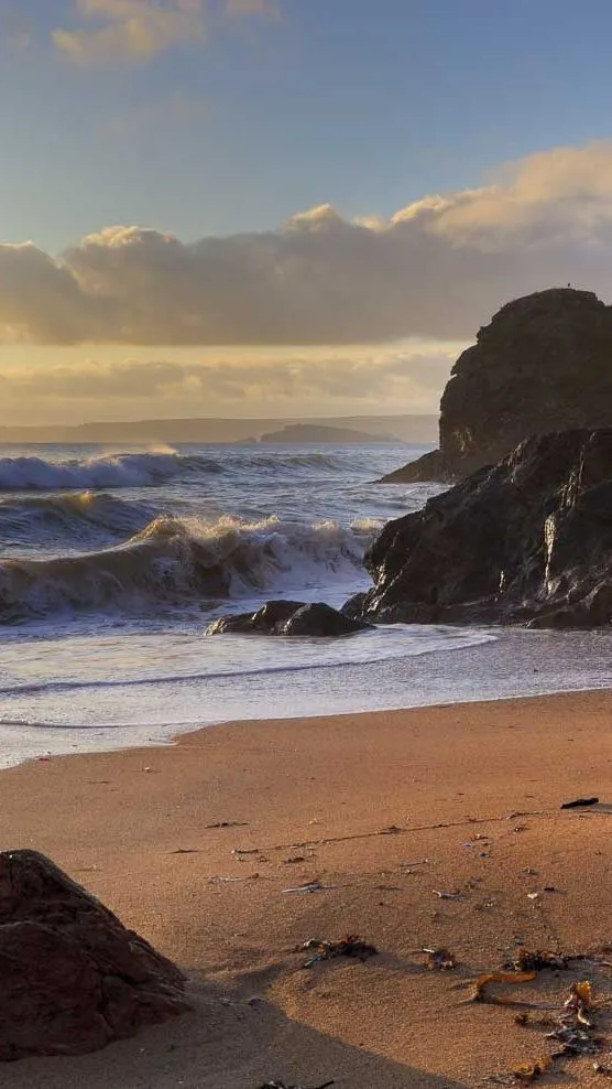 A scenic coastal view at sunset with waves crashing onto a sandy beach, surrounded by rugged cliffs and scattered rocks, bathed in golden light.