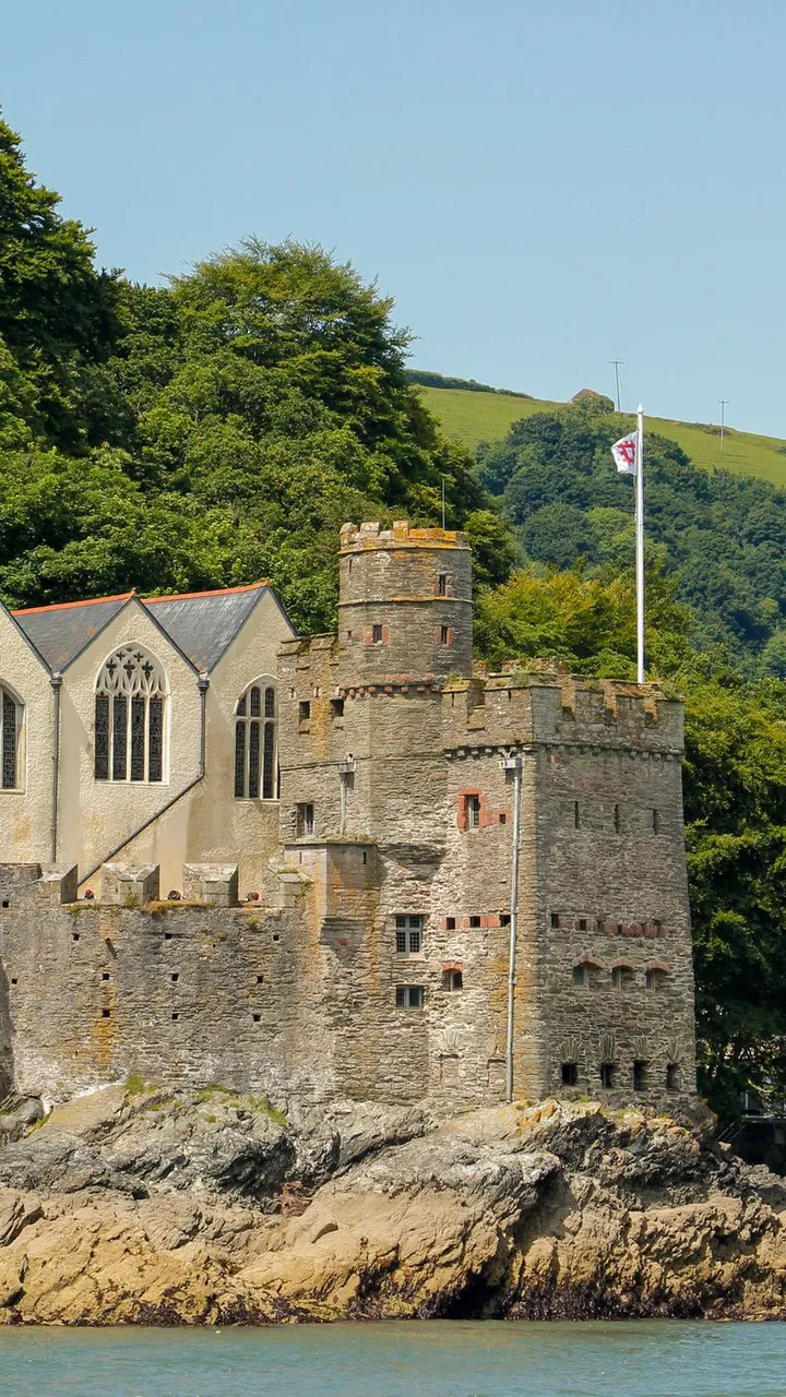 A historic stone castle with towers and arched windows sits on a rocky hillside by the water, surrounded by lush greenery, with flags flying under a clear blue sky.