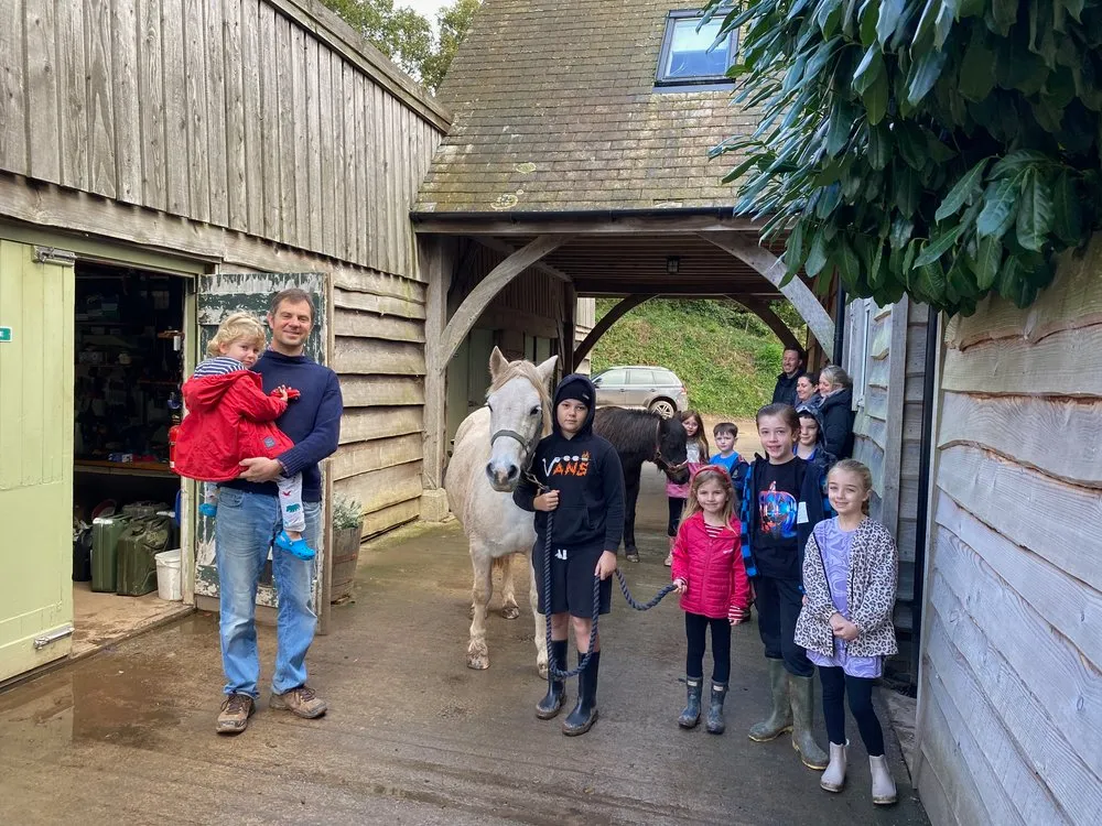 A group of children and adults stand outside a rustic wooden stable, posing with two ponies. A man holds a young child, while another child holds a pony's lead rope.