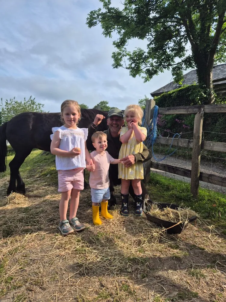 Three young children stand beside a smiling man near a black pony, holding fresh eggs.