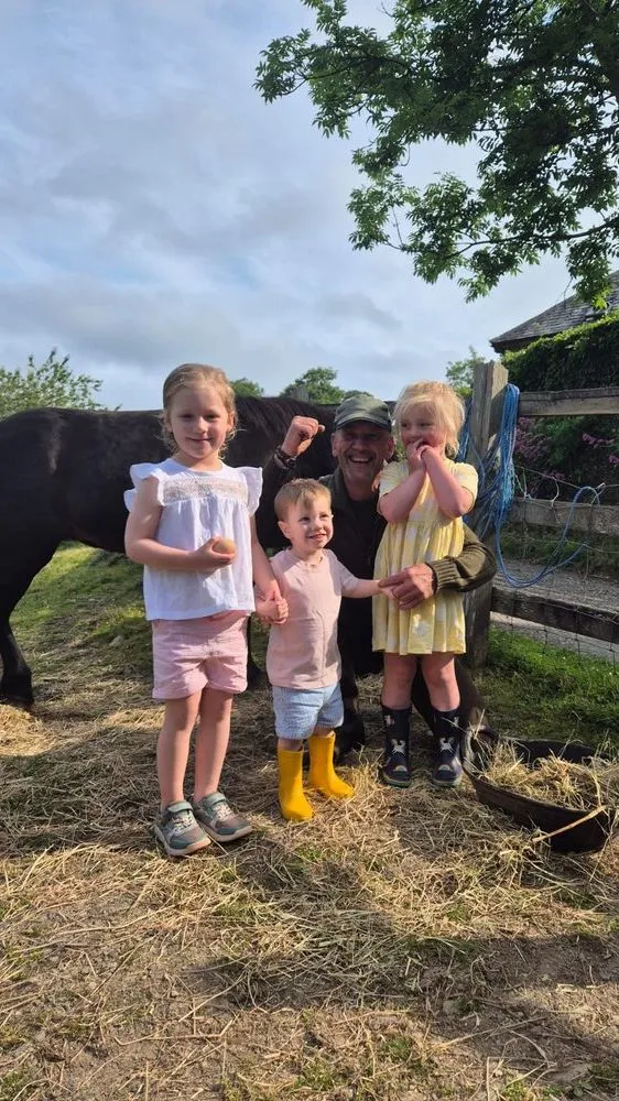 Three young children stand beside a smiling man near a black pony, holding fresh eggs.