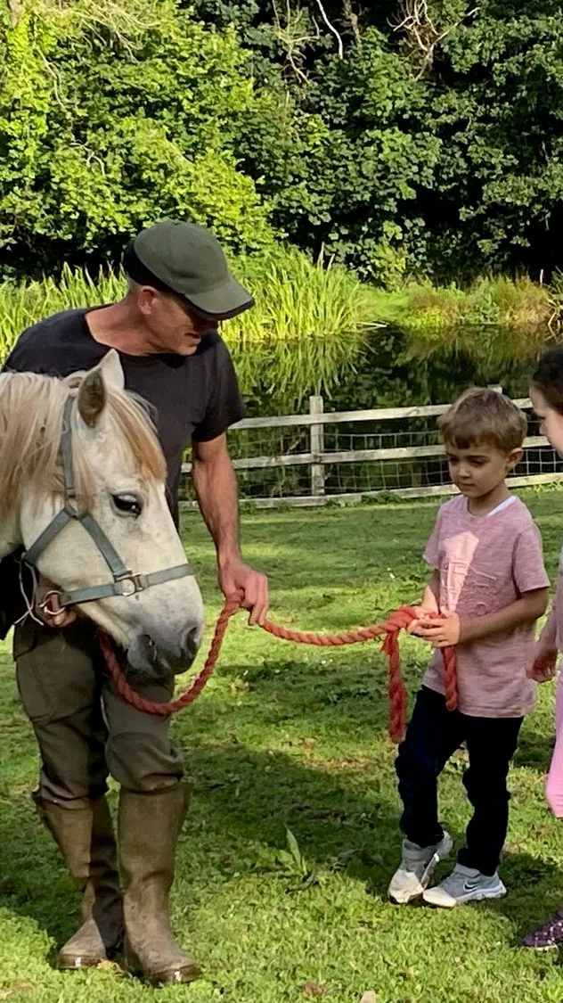 A man holds a red rope attached to a cream-colored horse while four young children stand nearby, watching attentively. Lush greenery and a pond are in the background.