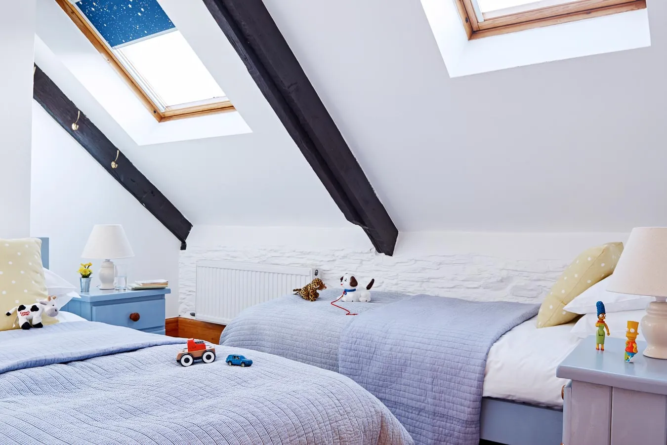 Cosy children's twin bedroom with sloped ceilings, skylights, soft lilac bedding, yellow polka dot cushions, wooden beams, and playful toys on the beds.