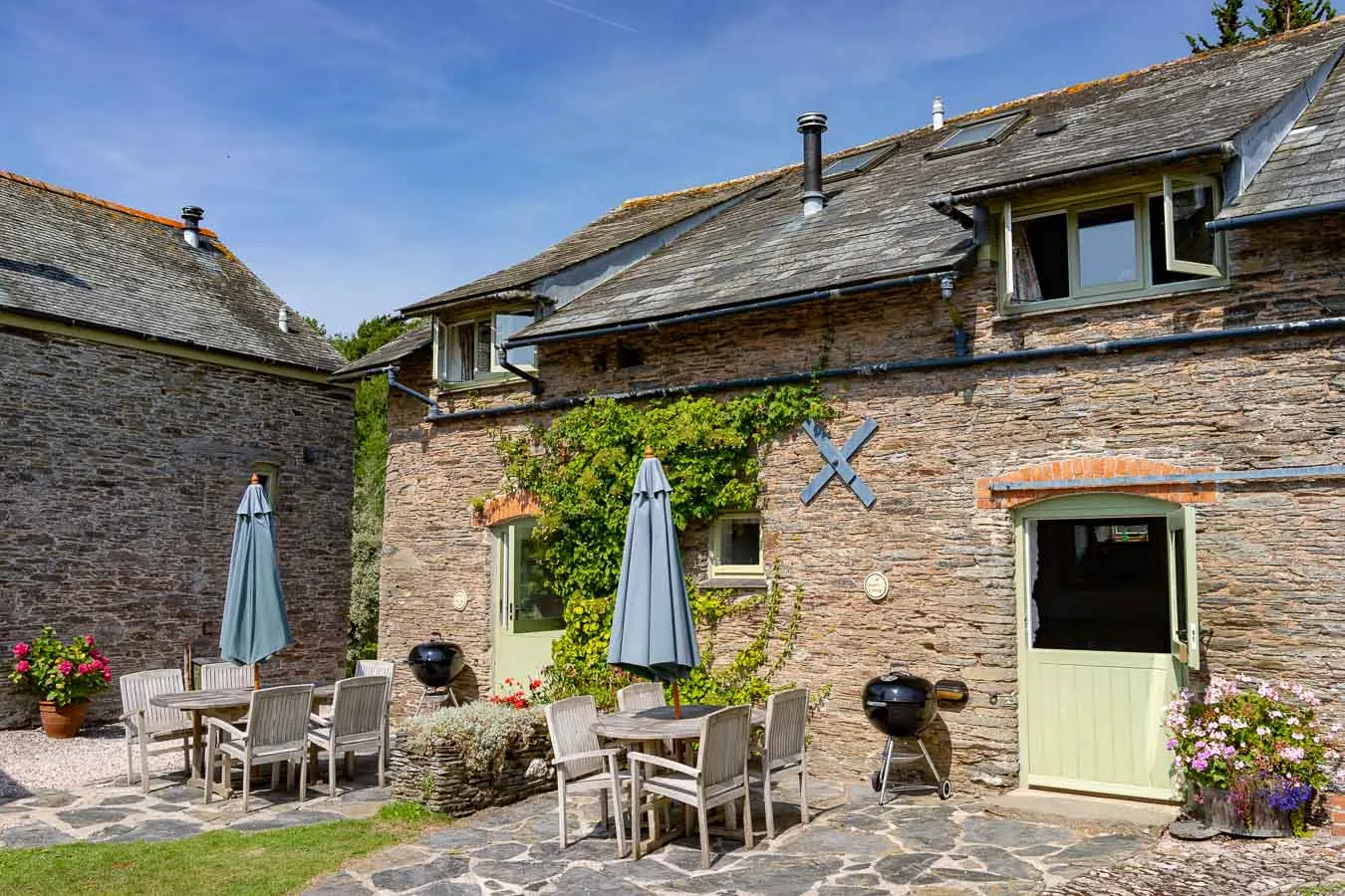 Charming stone cottage with ivy-covered walls, pastel green doors, outdoor dining tables with umbrellas, BBQ grills, and vibrant flower arrangements in a sunny courtyard.