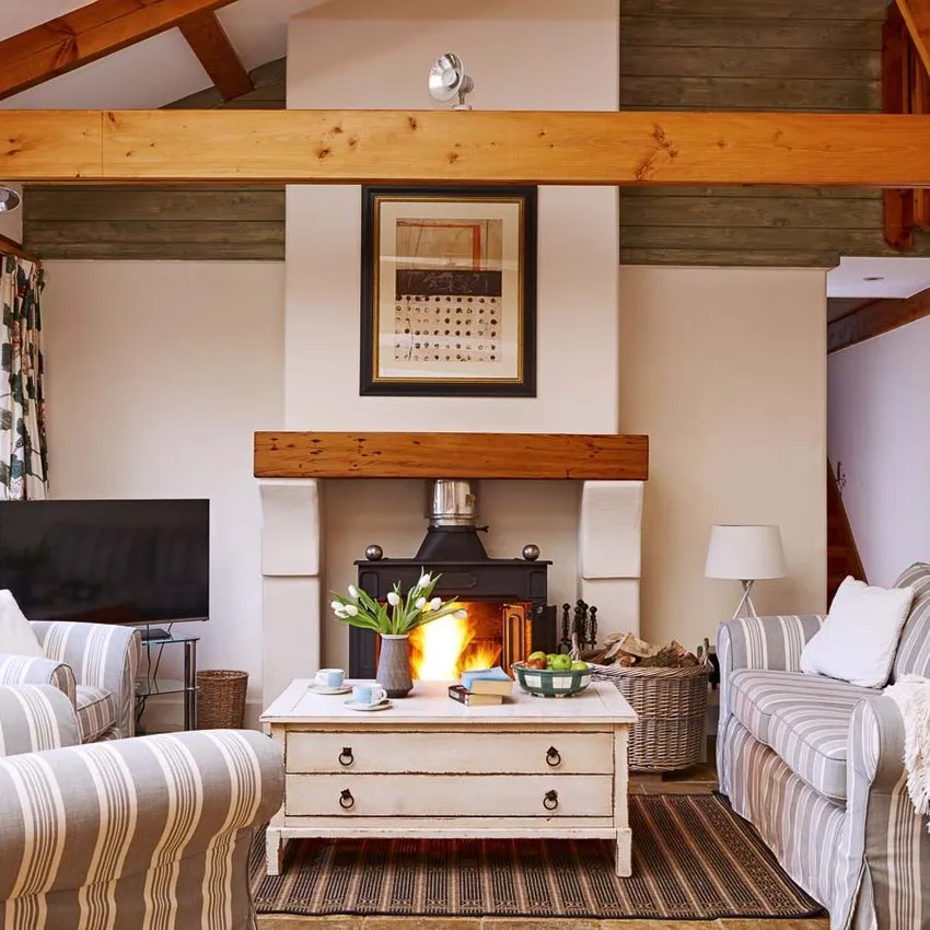 Cosy country-style living room with a wood-burning stove, striped sofas, wooden beams, and a rustic coffee table, creating a warm and inviting atmosphere.