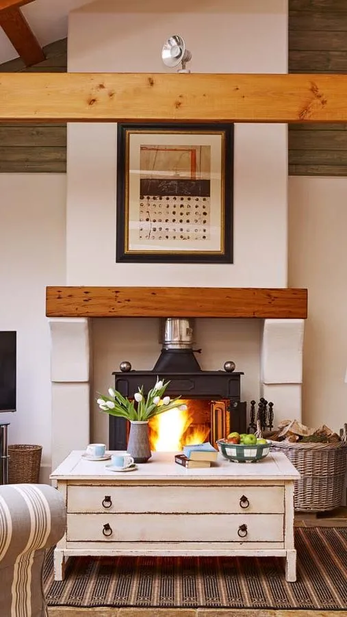 Cosy country-style living room with a wood-burning stove, striped sofas, wooden beams, and a rustic coffee table, creating a warm and inviting atmosphere.