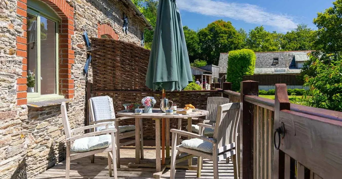 Charming outdoor deck with wooden furniture, a large green umbrella, and a rustic stone wall, set for a relaxing al fresco breakfast in the sunshine.